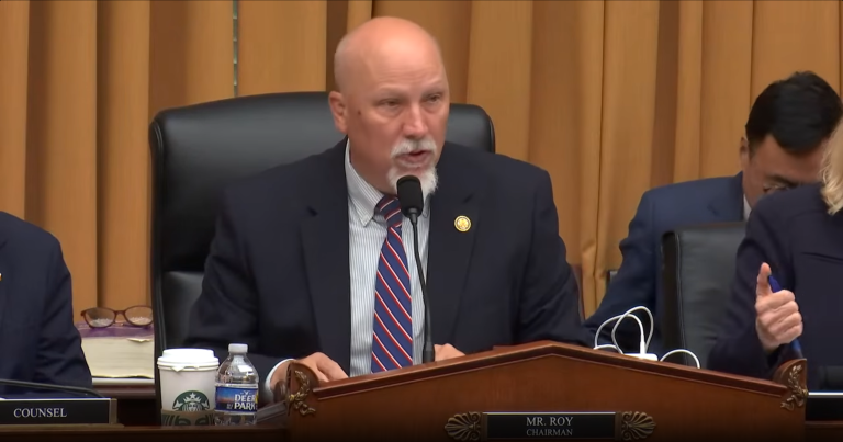 Chip Roy sits at the chairman’s desk during a congressional hearing, speaking into a microphone while wearing a dark suit and striped tie. Papers, a coffee cup, and water bottle sit nearby as staff members work in the background inside a formal House committee chamber.